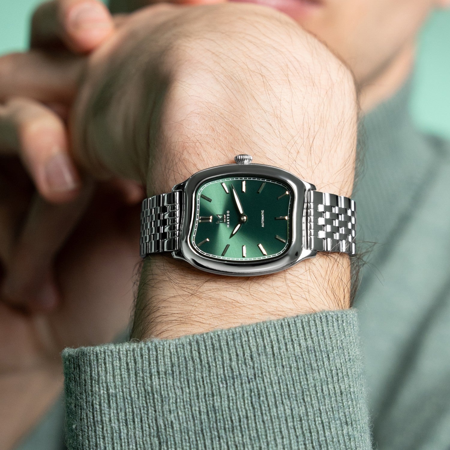 Green watch with silver case and band on a wrist, wearing a green shirt.