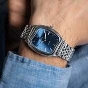 Silver watch with a blue face on a wrist, wearing a blue shirt.